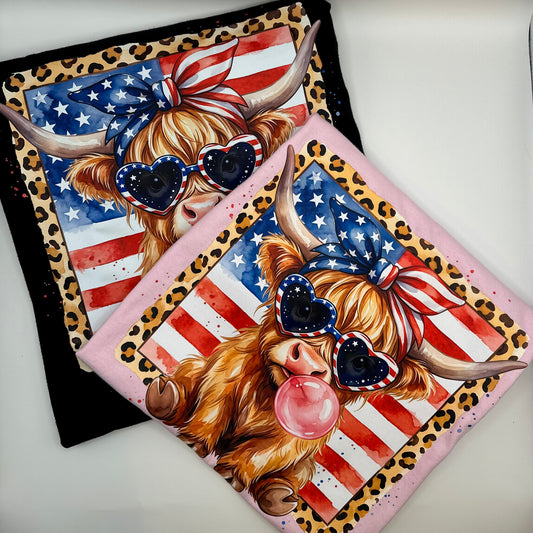 Two T-shirts folded into squares with printed cartoon Highland cows wearing heart-shaped sunglasses, a US flag bandana, and a leopard-print border, shown overlapping on a white background