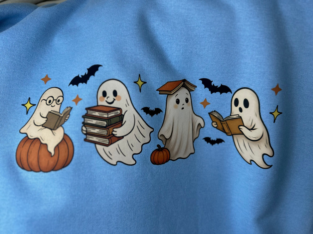 A close-up of a light blue fabric featuring a printed Halloween graphic of four cute ghosts reading books, with pumpkins, bats, and stars.