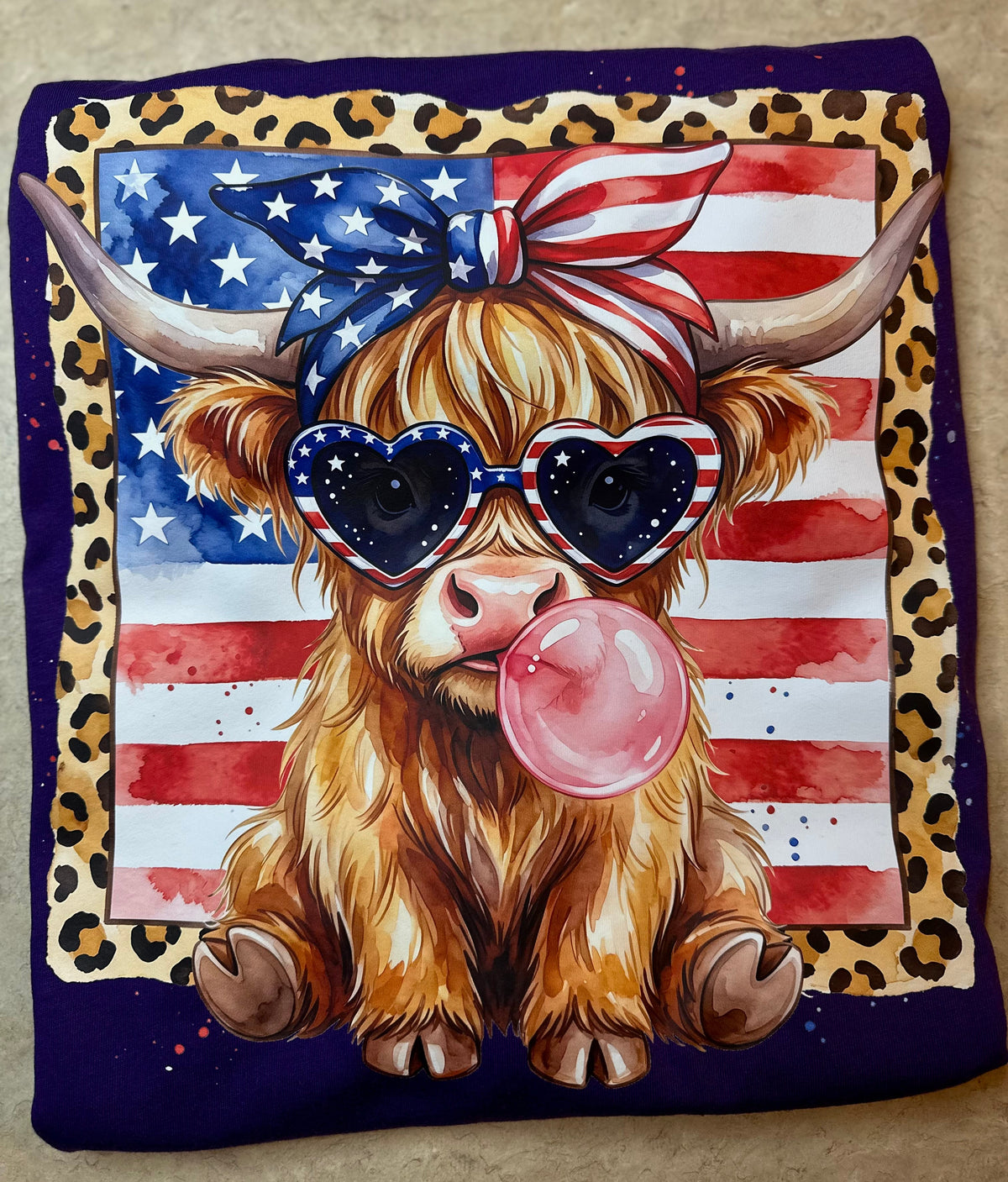 A T-shirt folded into a square featuring a colorful printed design of a fluffy Highland cow wearing heart-shaped sunglasses, a red-and-white bandana, and a pink bubblegum bubble, set against an American flag and leopard-print border on a dark purple shirt.