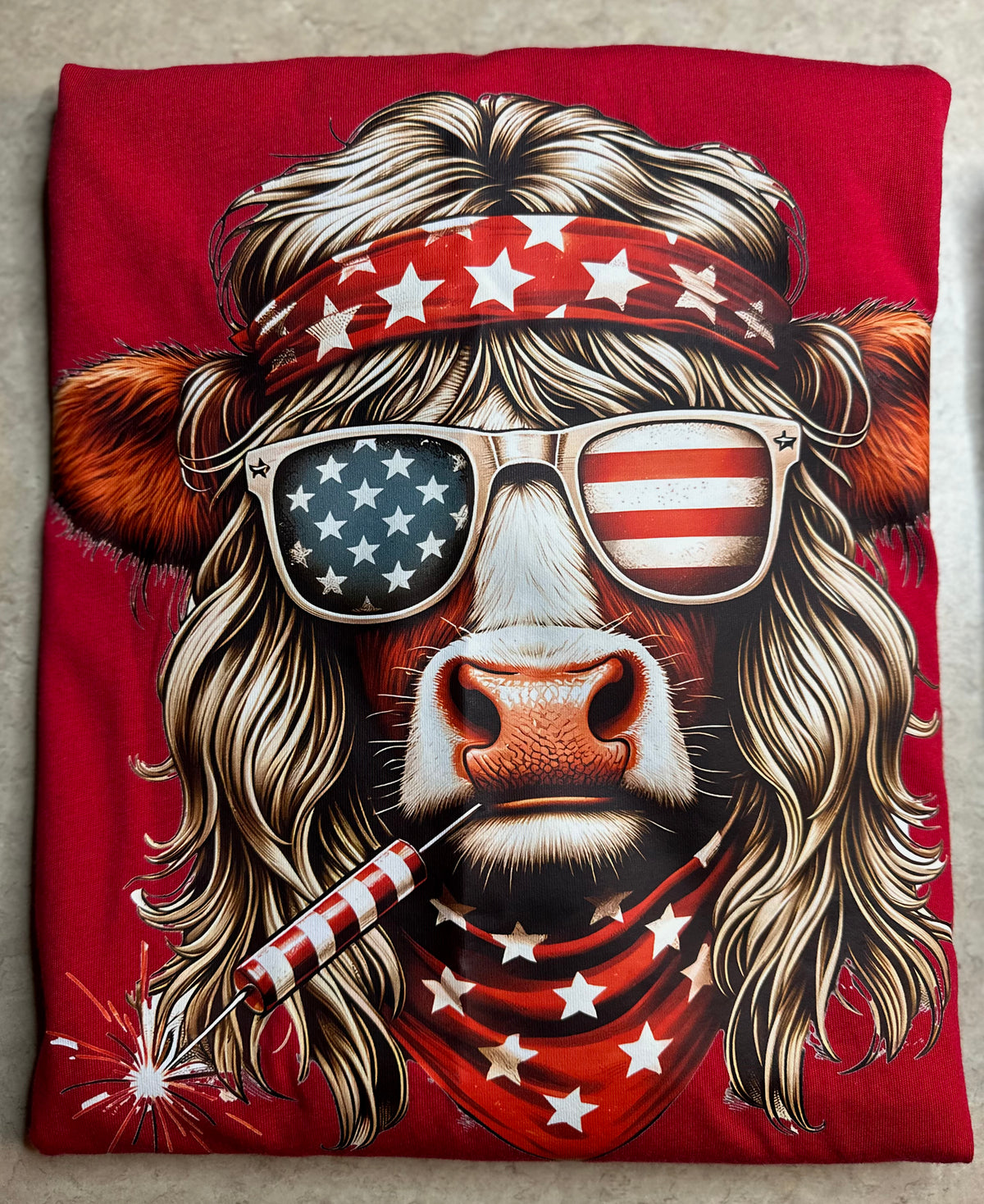 A folded red T-shirt featuring a large graphic print of a cow with long blonde hair, a red bandana with white stars, star-spangled sunglasses, and a patriotic American theme.