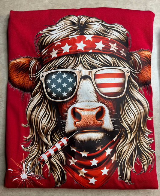 A folded red T-shirt featuring a large graphic print of a cow with long blonde hair, a red bandana with white stars, star-spangled sunglasses, and a patriotic American theme.
