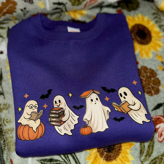 A folded purple sweatshirt featuring a cartoon Halloween graphic of four ghosts reading, sitting on pumpkins, with bats and stars, displayed on a floral blanket.